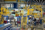 In a large, brightly-lit facility with blue- and yellow-colored scaffolding, military aircraft are lined up in varying states of assembly.