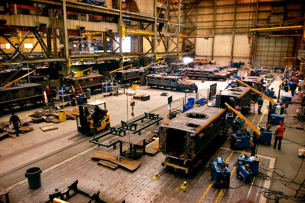 Inside a large manufacturing facility, multiple military vehicles are in various states of assembly.