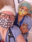 two white females in blue scrubs wearing masks, head coverings and stethoscopes.
