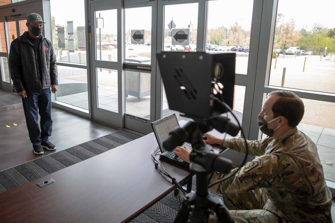 A soldier sits facing a camera, which is pointed at a man, standing, in civilian clothing about six feet away.