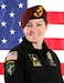 White female in black uniform with patches and maroon beret with gold and black flash stands in front of an American flag.