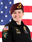 White female in black uniform with patches and maroon beret with gold and black flash stands in front of an American flag.