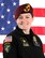 White female in black uniform with patches and maroon beret with gold and black flash stands in front of an American flag.