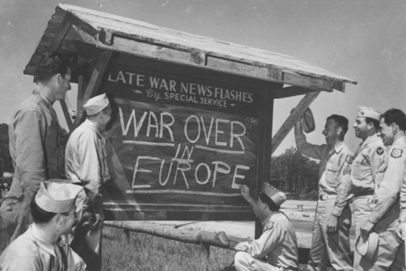 Service members stand around a sign reading "War Over in Europe"