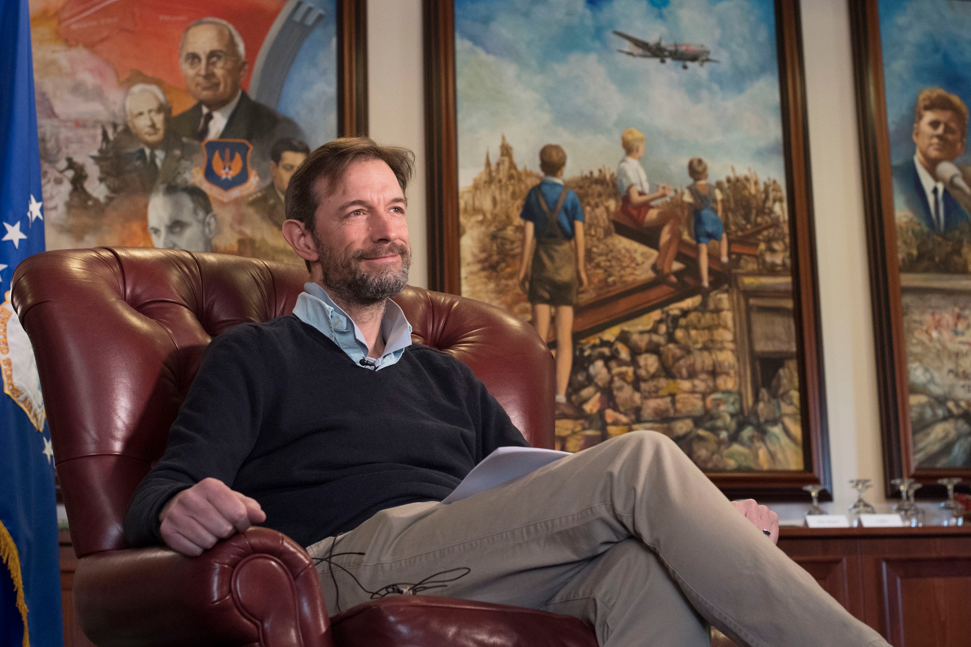 A man sits in a chair with historical paintings in the background.