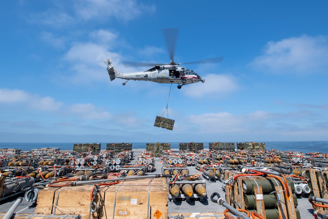 A helicopter uses a cable to lift a large container from a military ship.