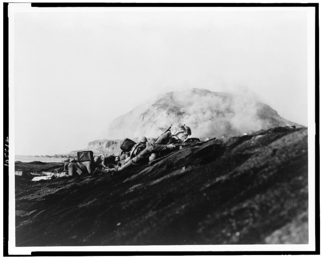 Marines lay prone with guns pointed on a black hill. A mountain rises in the background.