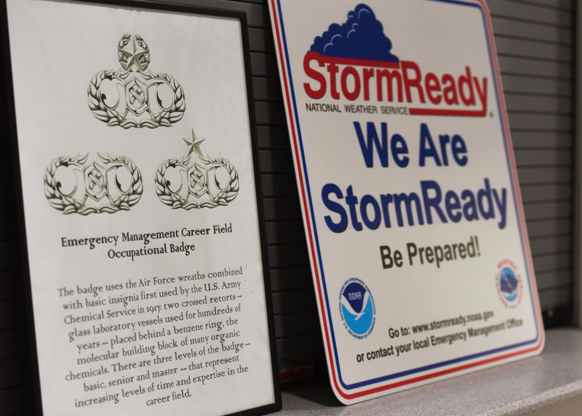 Signs in the 509th Bomb Wing Emergency Management work center display the Career Field Occupational Badge history and National Weather Service 'Storm Ready' information.  The badge symbols and adverse weather sign show the range of different Emergency Management responsibilities.