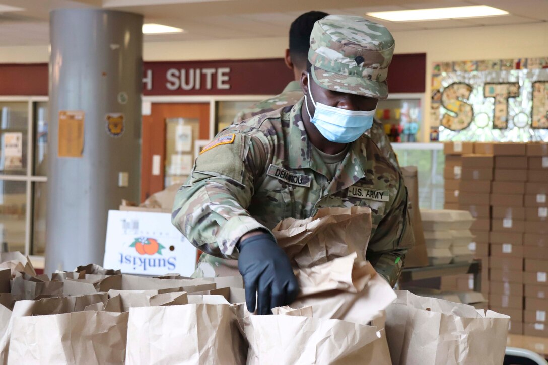 A soldier wearing a face mask and gloves packs meals into paper bags.