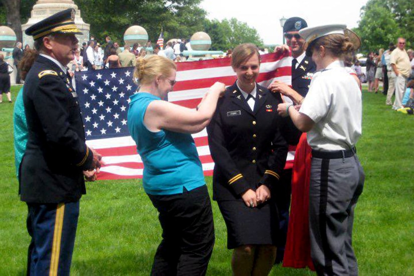 A soldier stands smiling around her family as they affix decorations to her uniform.