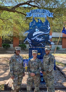 Three females in green camouflage uniforms stand a blue sign outside of a building.