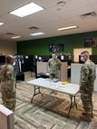 Three males in green camouflage uniforms and face masks stand around a table in a room with green and grey walls and grey chevron carpets.