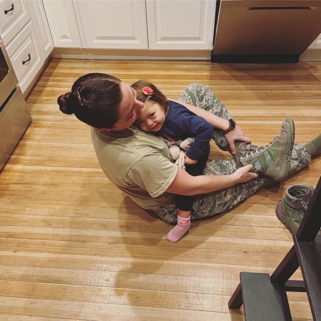 Mother and daughter hug sitting on the floor while mother puts boots on for work.