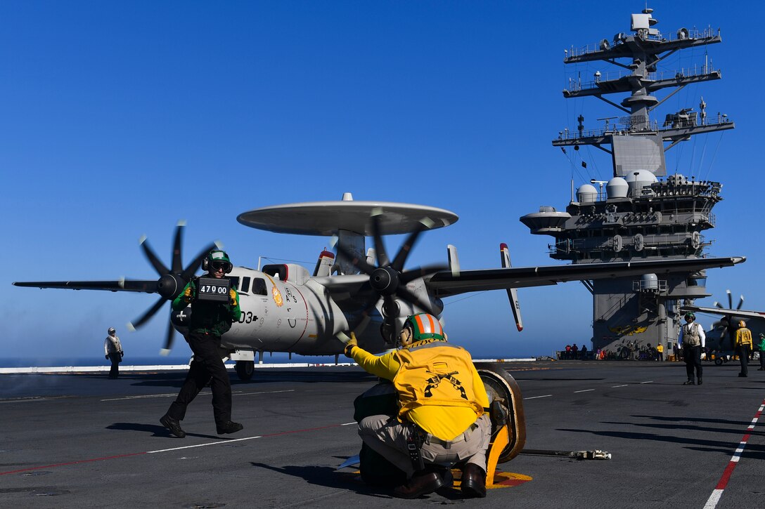 A sailor signals a military aircraft on the deck of an aircraft carrier.