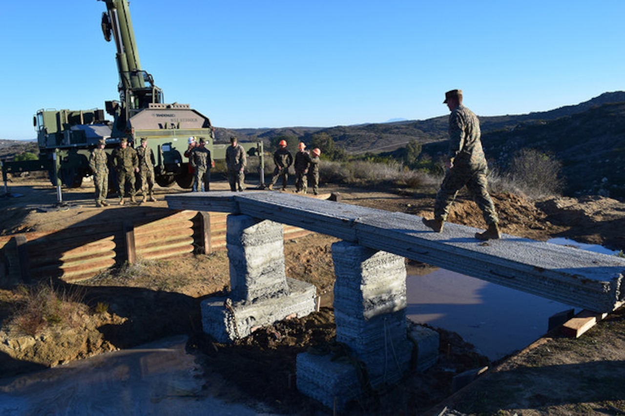 A service member crosses a footbridge over a small stream.