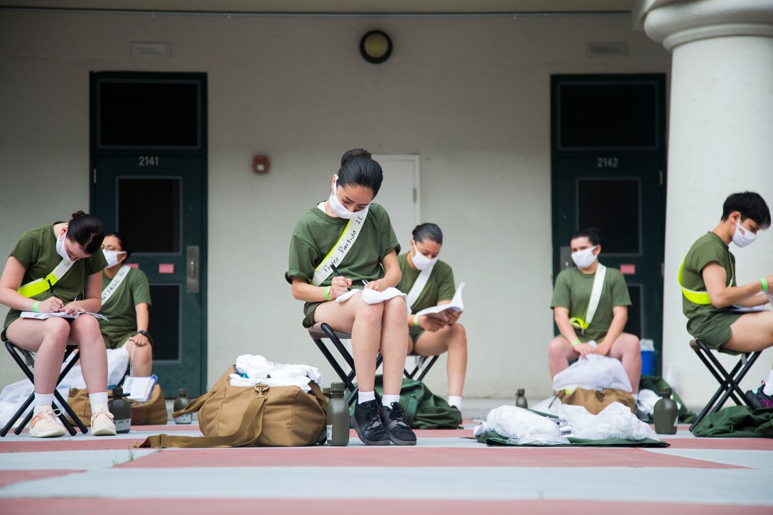 Marine Corps recruits wearing face masks sit on chairs while writing on notepads.