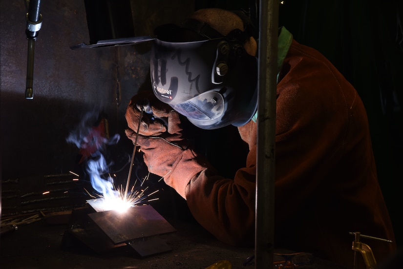 Sparks fly as an airman wearing a helmet and mask welds metal.