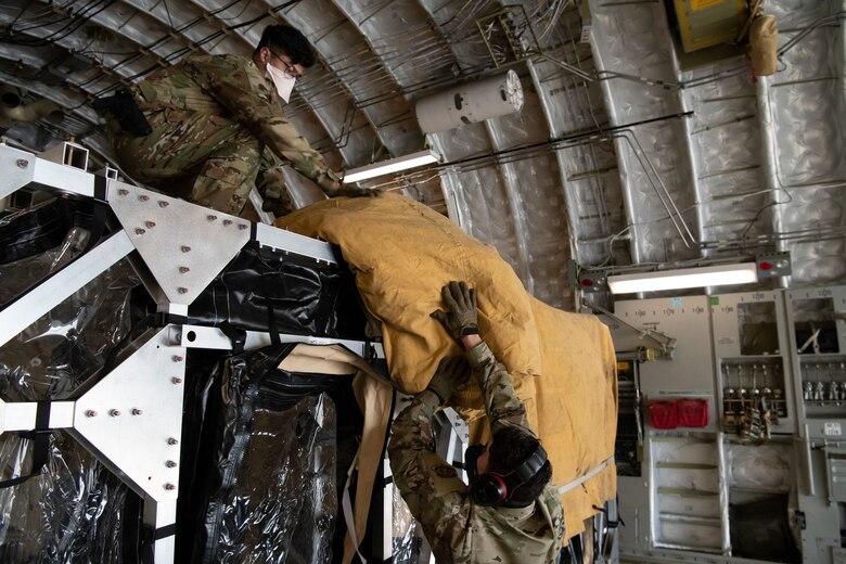 Senior Master Sgt. Andrew Silkworth, 436th Aerial Port Squadron airfreight superintendent, throws a tarp up to Airman 1st Class Jose Gomez, 14th Airlift Squadron loadmaster, at Dover Air Force Base, Delaware, April 30, 2020. The tarps were placed on top of the newly arrived Transport Isolation Systems due to inclement weather at Dover AFB. In accordance with health protection policies, Dover AFB will serve as the sole hub for TIS decontamination on the East Coast. (U.S. Air Force photo by Senior Airman Christopher Quail)