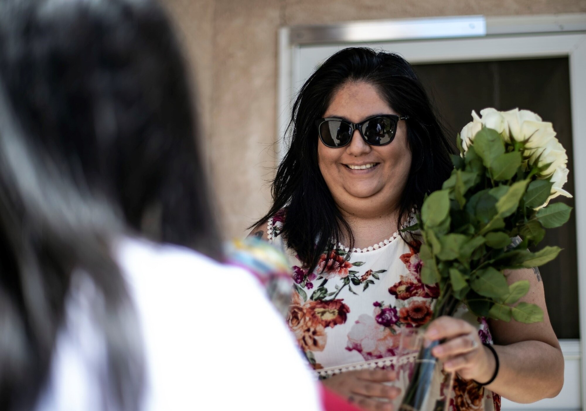 A smiling woman in sunglasses looks at another woman and holds a bouquet of flowers.