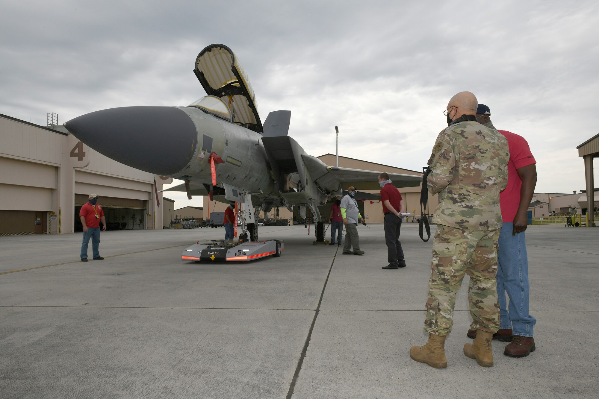 Photo shows a group of people around an F-15 aircraft which has a machine hooked up to the front wheel to direct the aircraft into place.