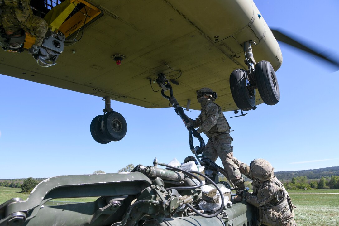 A soldier stands atop a large weapon and hooks it up to the bottom of a helicopter using a sling.