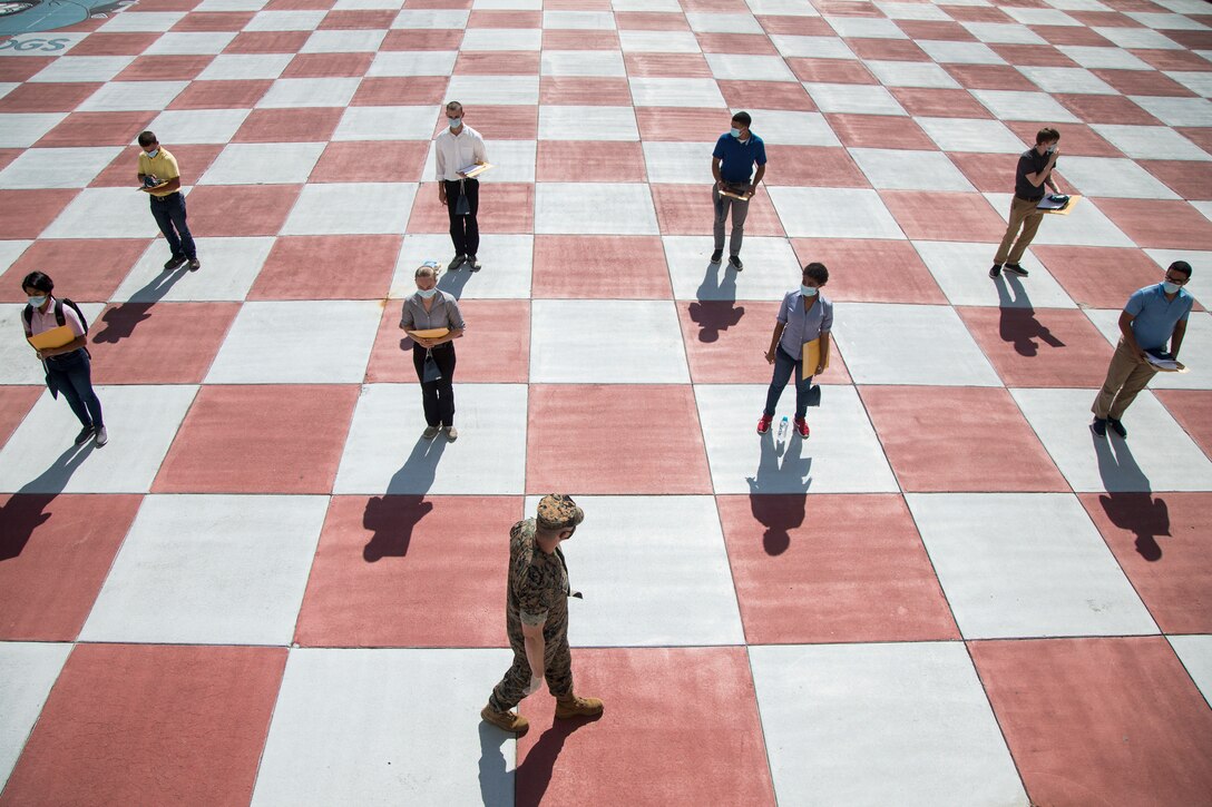 People stand in two rows on a large checkerboard floor as a Marine walks in front of them.