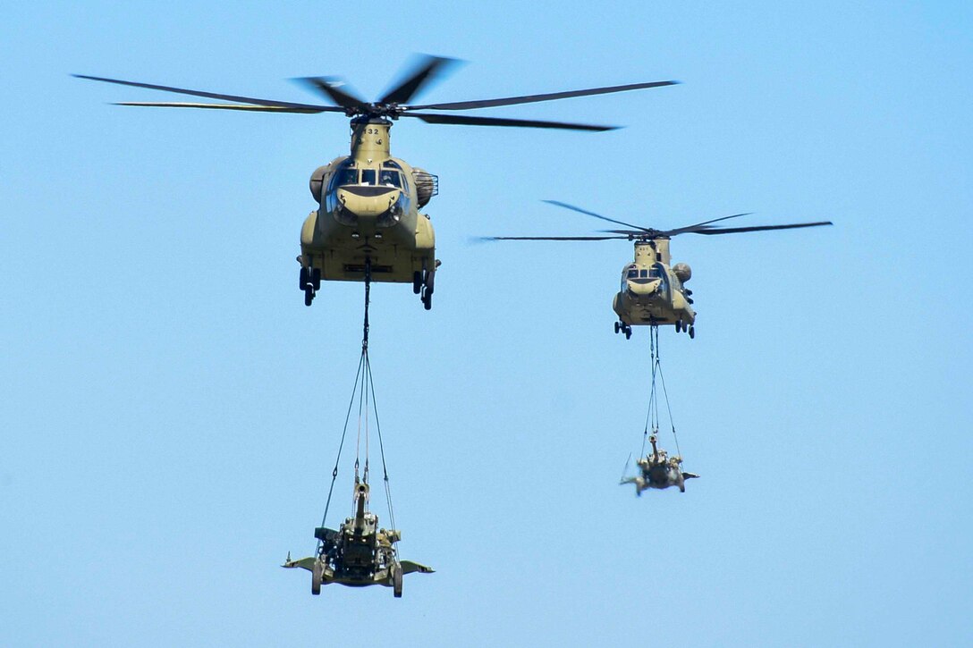 Two Army helicopters fly with howitzers suspended below them by ropes.