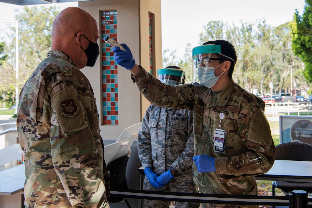 An airman checks the temperature of a senior officer as another service member watches.