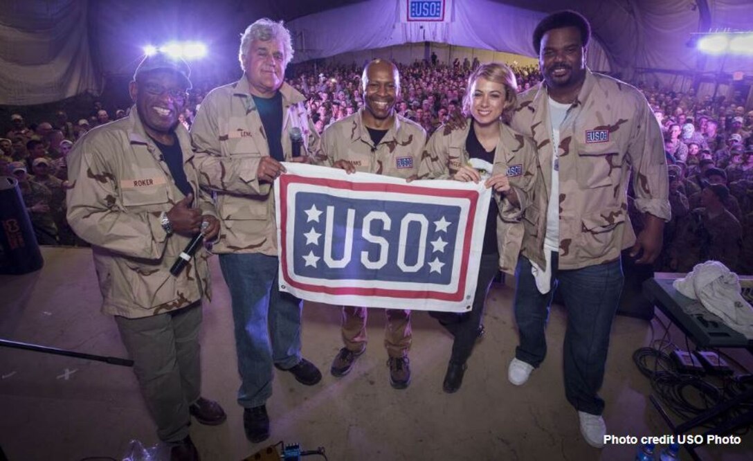 Four men and one woman hold a USO flag with hundreds of troops in the background.