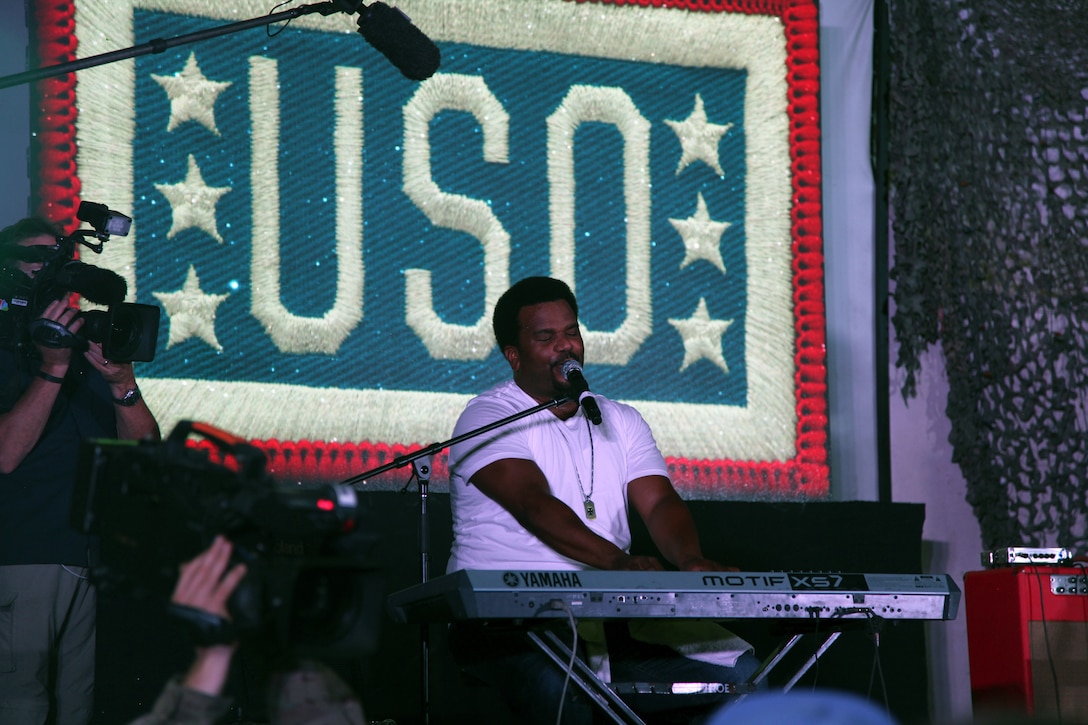 A man plays a keyboard and sings in front of a giant screen that says “USO.”