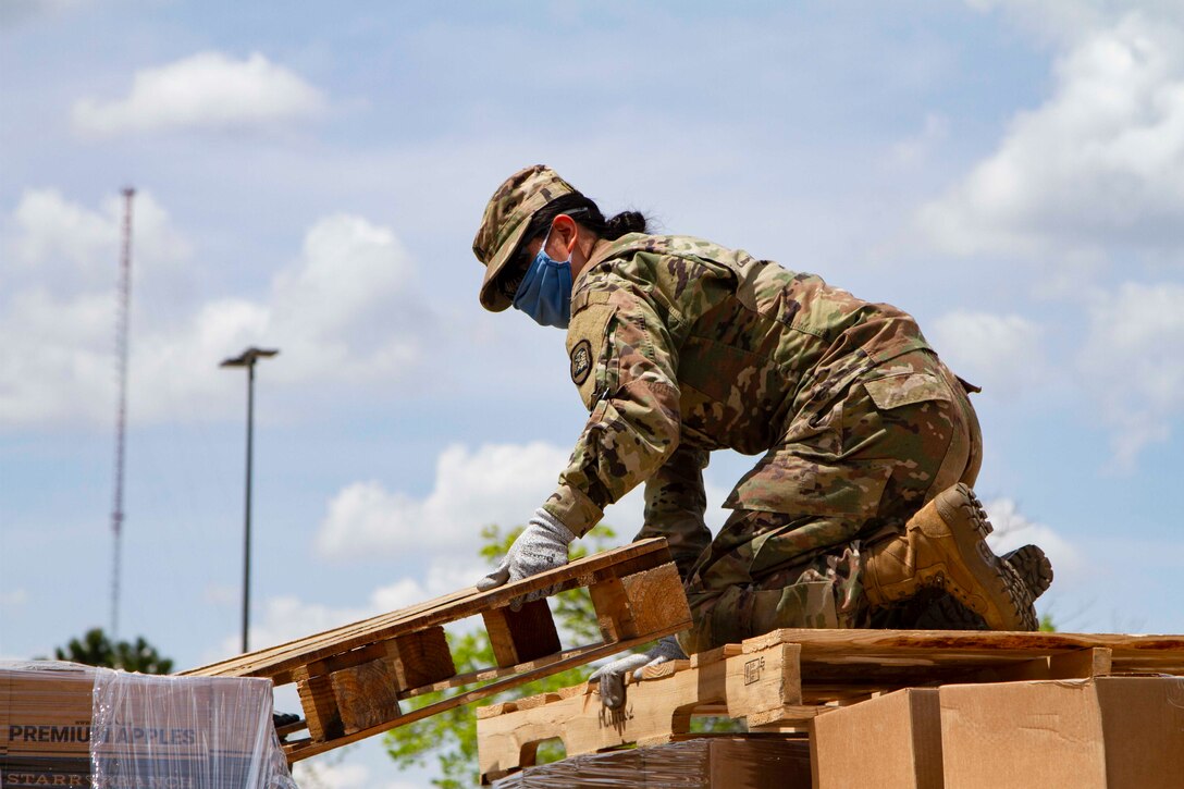 A soldier wearing a face mask kneels on pallets while unloading.