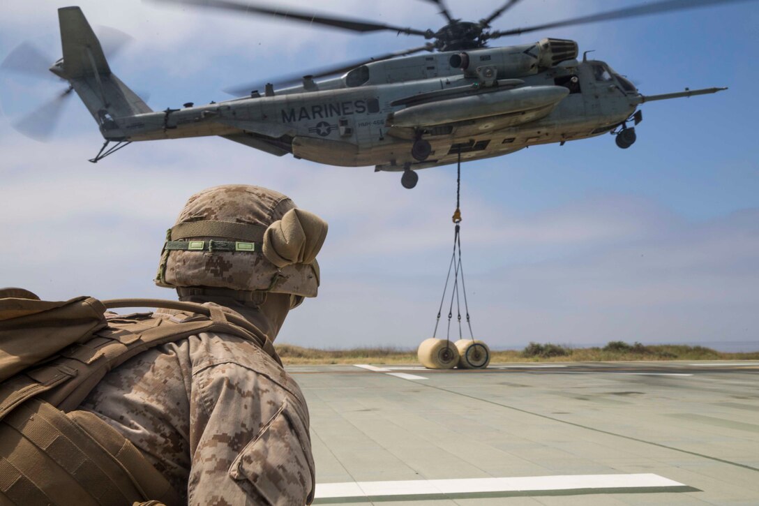 A Marine looks on as a Marine Corps helicopter hovers with cargo hanging from rope beneath it.