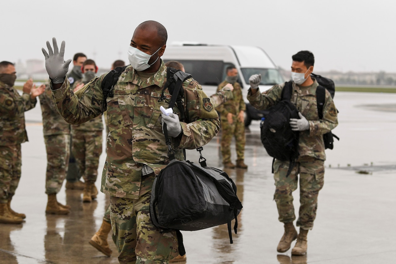 Airmen wearing face masks and carrying bags walk on a paved lot and exchange greetings with fellow troops.