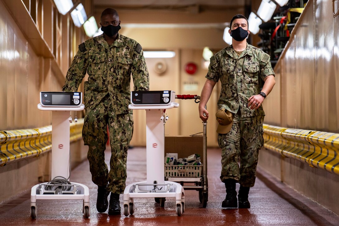 Two sailors wearing protective face masks walk down the hall transporting medical equipment.