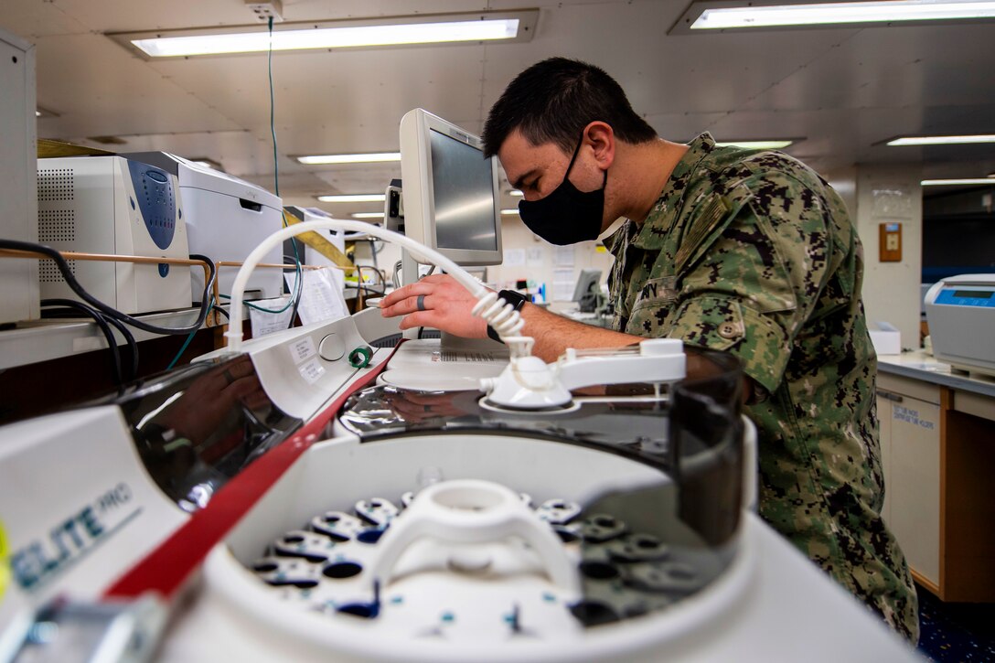 A sailor wearing a protective face mask troubleshoots medical equipment.