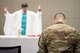 Chaplain holds hands in the air in front of alter next to Soldier with his head bowed