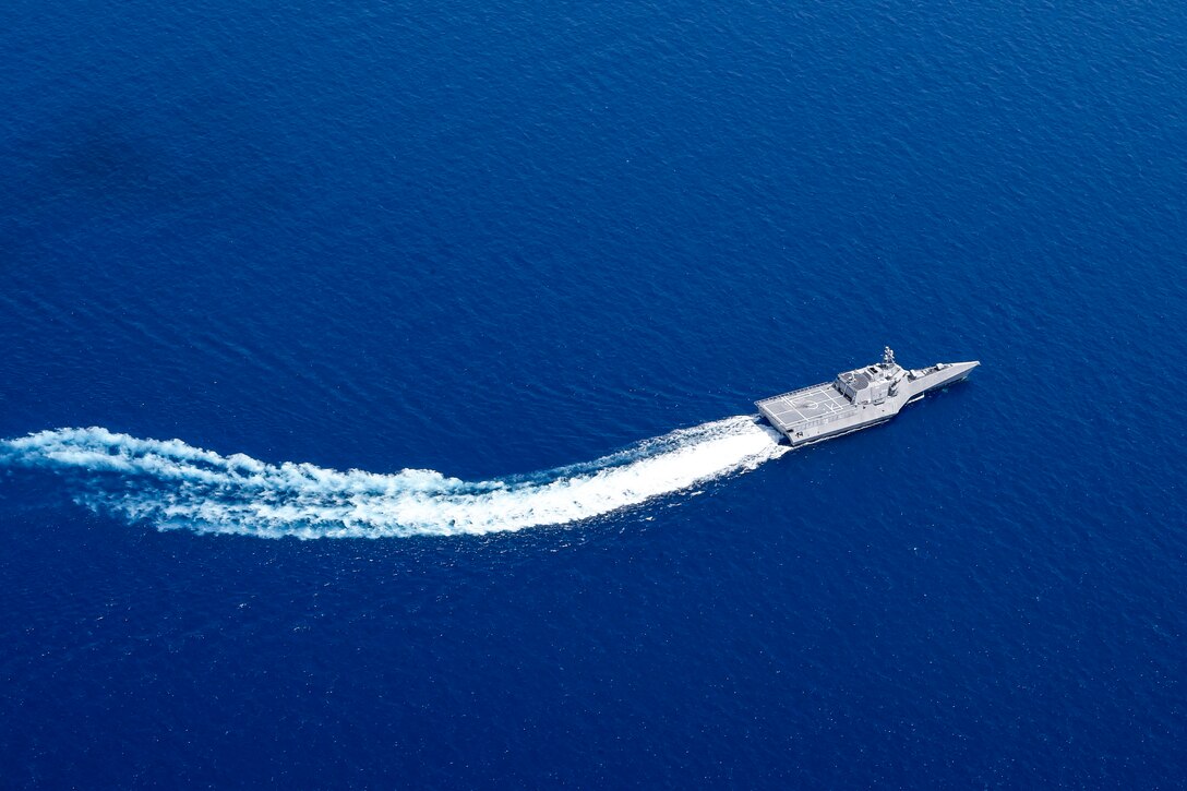 A ship, seen from overhead, leaves a sweeping white wake as it travels in vivid blue waters.