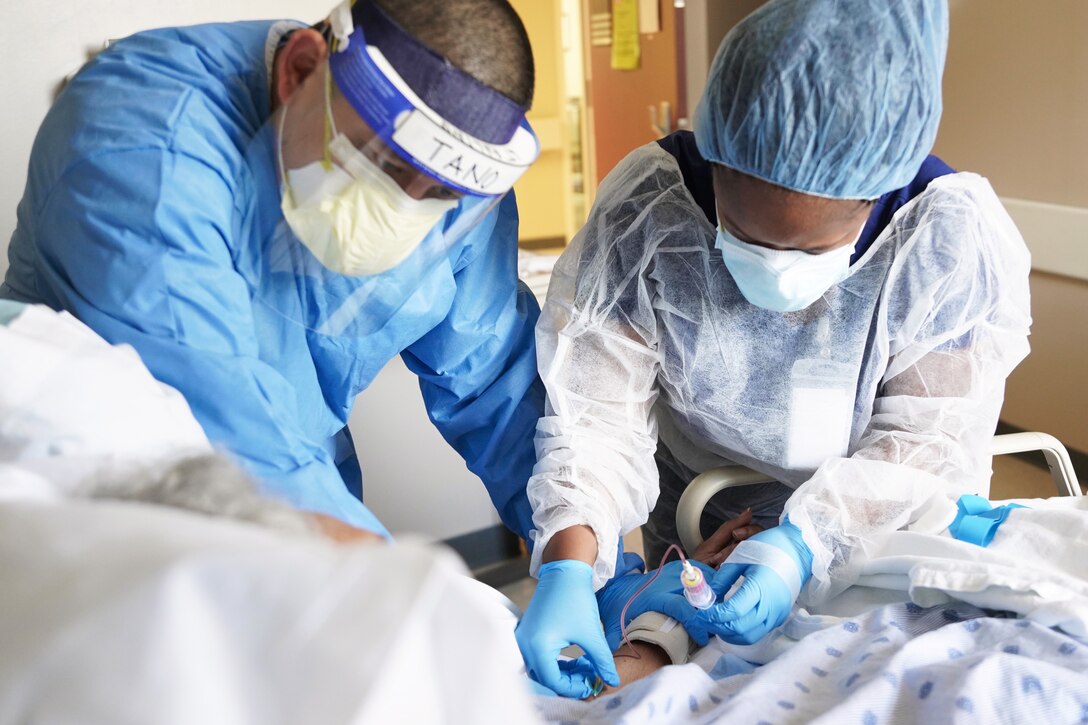 A Navy sailor and a technician wearing protective face masks and gowns draws blood from a patient