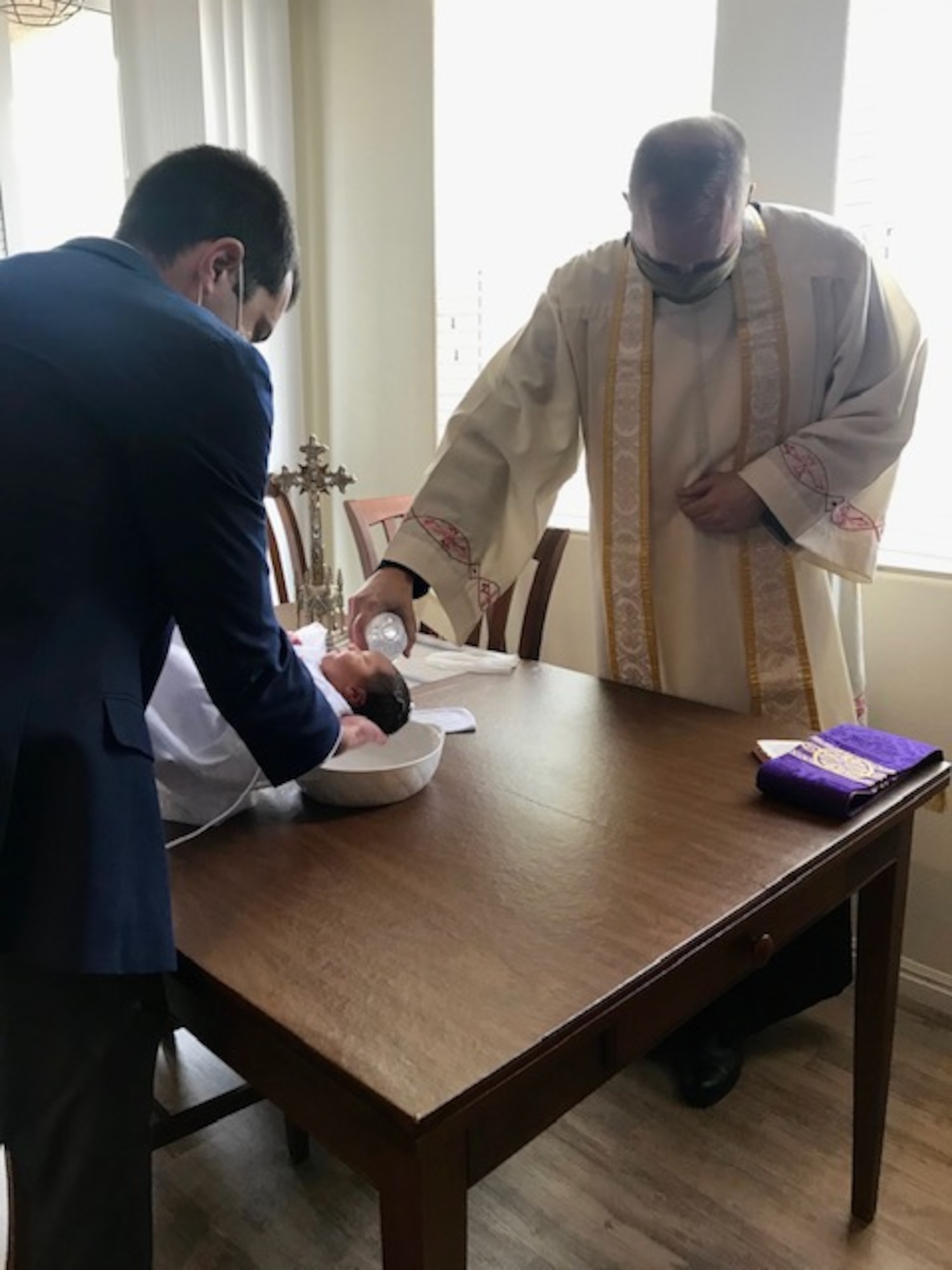 A Catholic priest in white robes and a facemask, baptizes an infant with water as the father holds the infant.