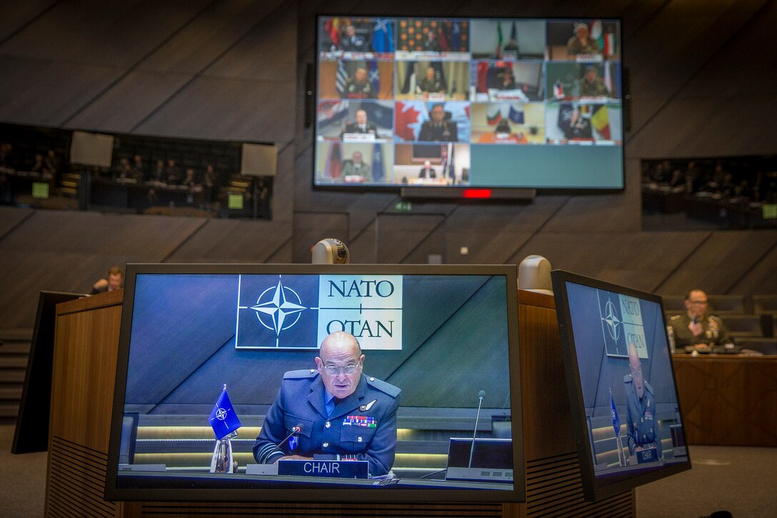Multiple video monitors in a council room display men in military uniforms on a screen.