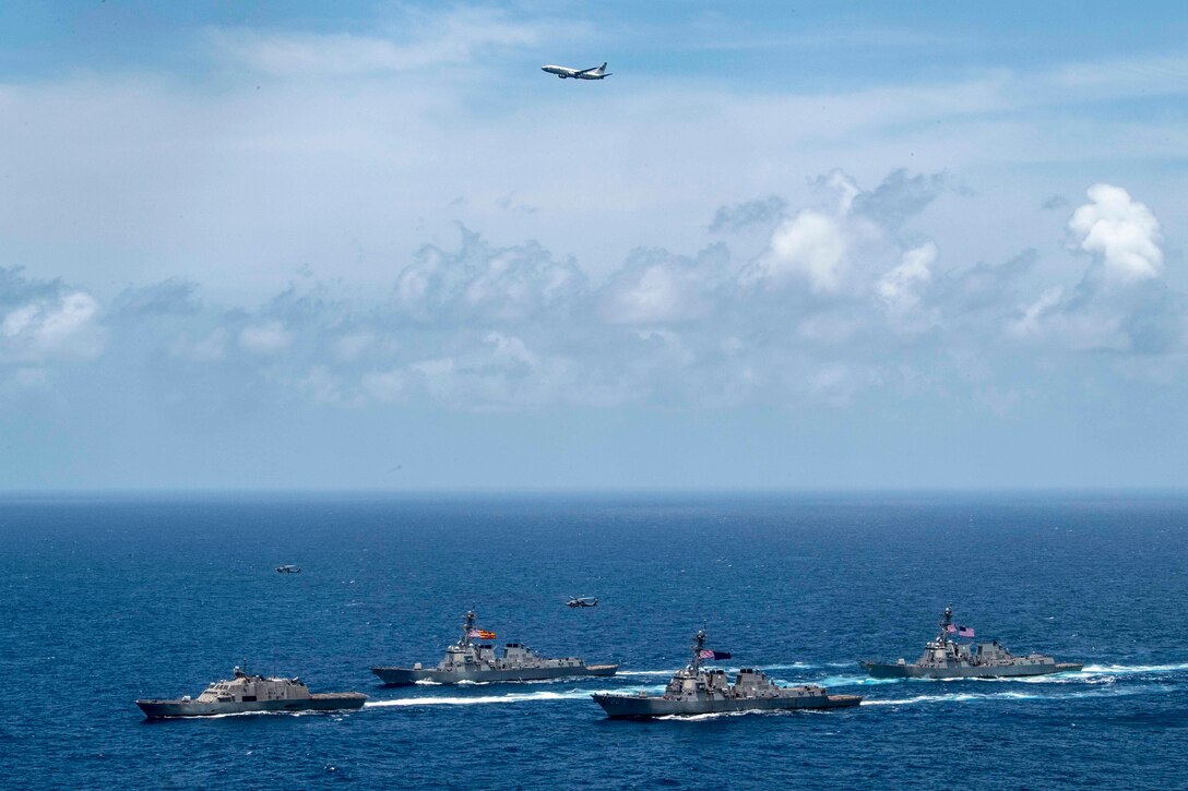 Four ships travel in formation as an aircraft flies above them.