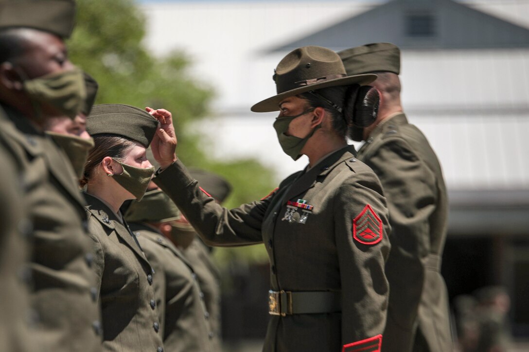 A Marine Corps drill sergeant in a mask adjusts the cover of another Marine.