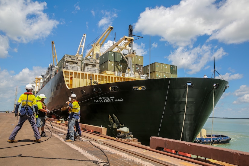 Four men pull on a cable attached to a massive container ship at port.