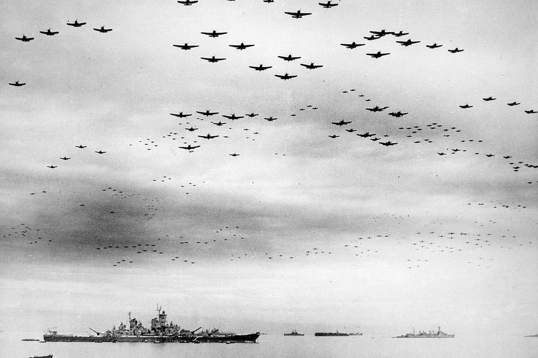 Hundreds of World War II-era aircraft fly over a large ship.