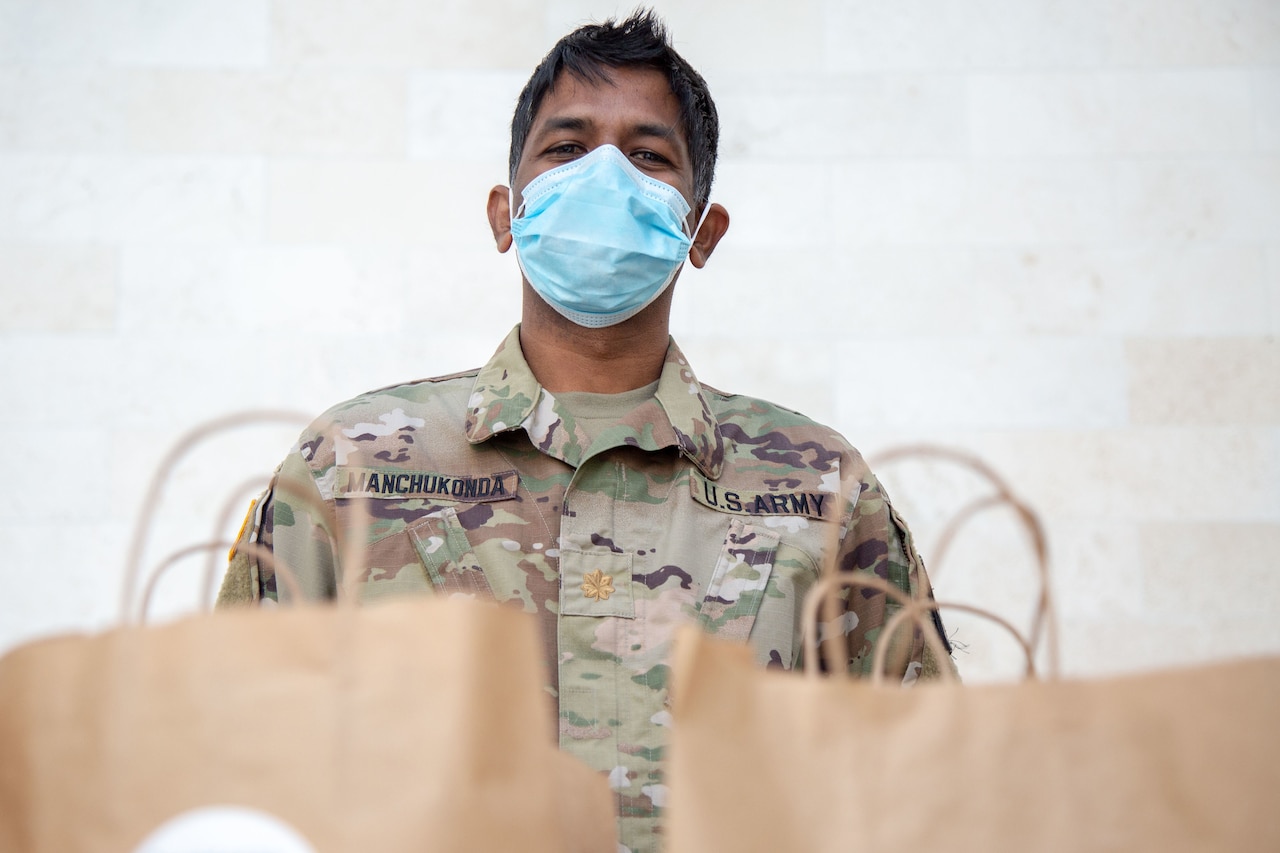 A man in a military uniform and face mask stands ready to distribute food packed in shopping bags.