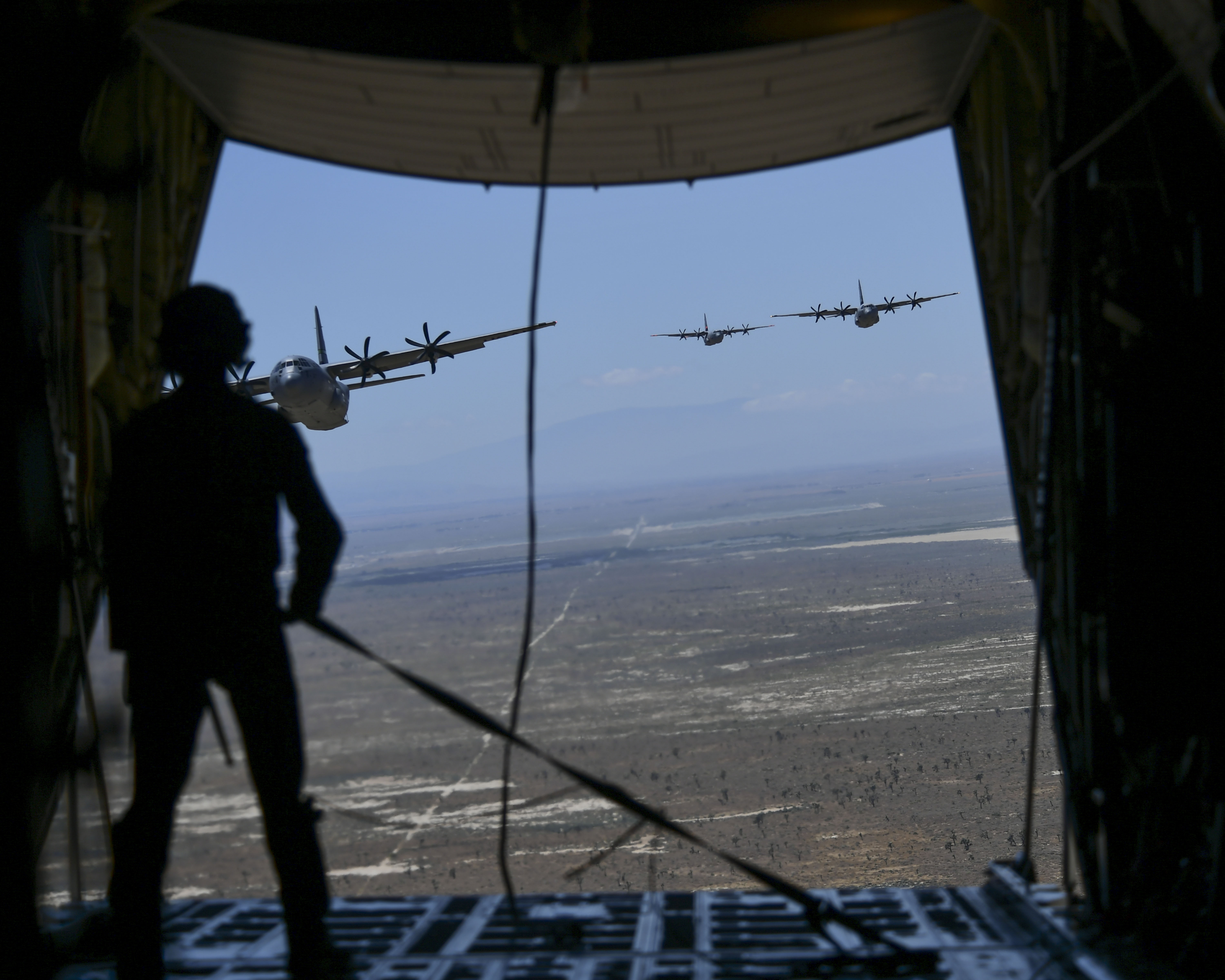 A California Air National Guard loadmaster is seen holding a tie down strap inside the back of a military California Air National Guard C-130J Super Hercules aircraft. In front of him the back of the aircraft's ramp is open to show three additional C-130J Super Hercules aircraft from the California Air National Guard following in close pursuit at a lower altitude above a drop zone in the Palmdale desert.