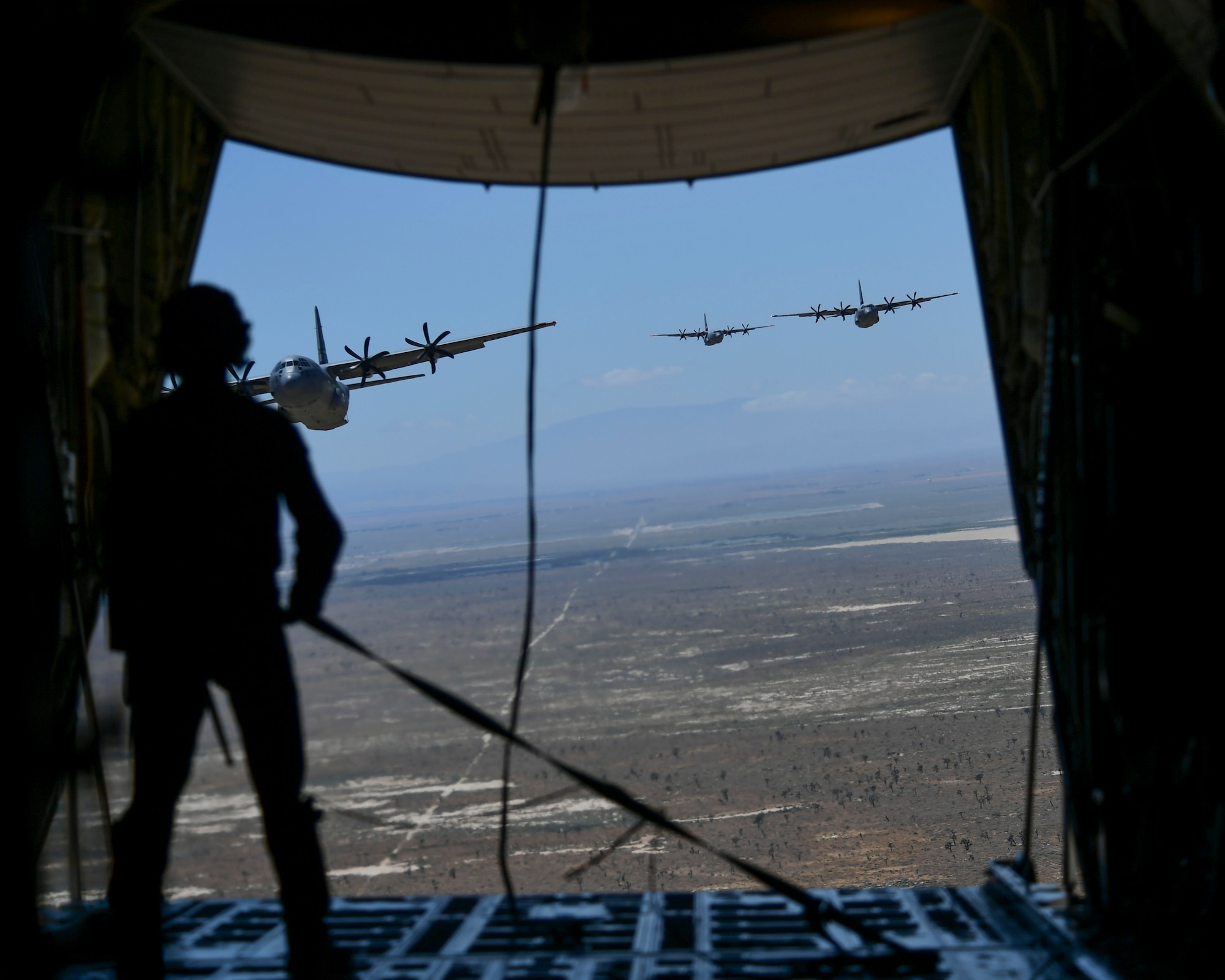 A California Air National Guard loadmaster is seen holding a tie down strap inside the back of a military California Air National Guard C-130J Super Hercules aircraft. In front of him the back of the aircraft's ramp is open to show three additional C-130J Super Hercules aircraft from the California Air National Guard following in close pursuit at a lower altitude above a drop zone in the Palmdale desert.