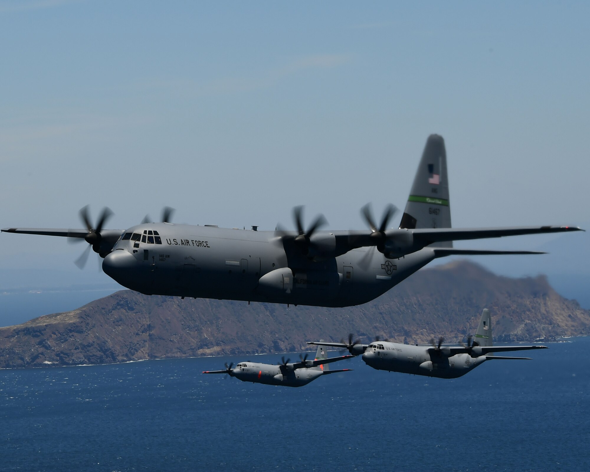 Three military California Air National Guard C-130J aircraft fly in close formation over the blue Pacific Ocean with the Anacapa Islands in the background.