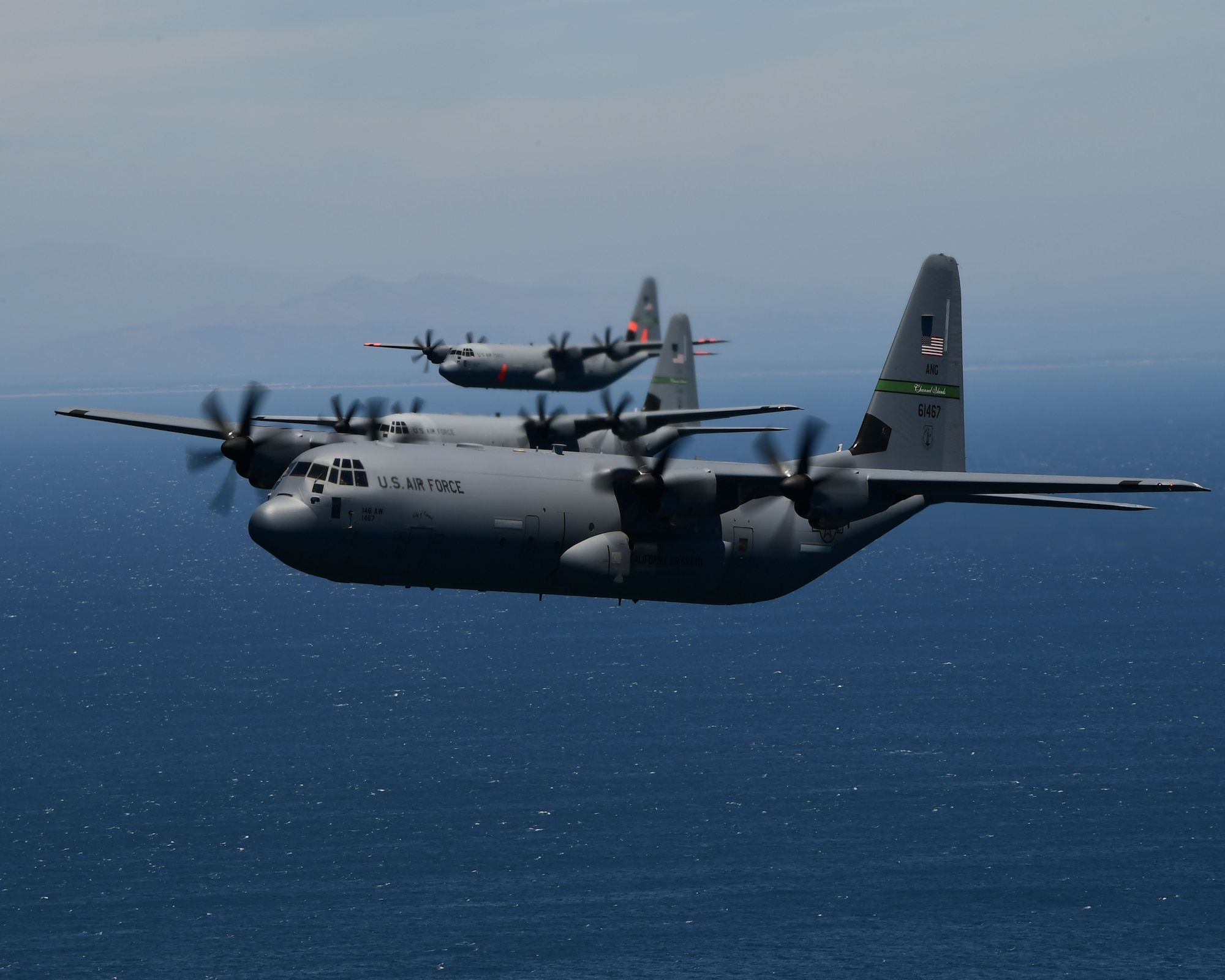 Three military California Air National Guard C-130J aircraft fly in close formation over the blue Pacific Ocean.