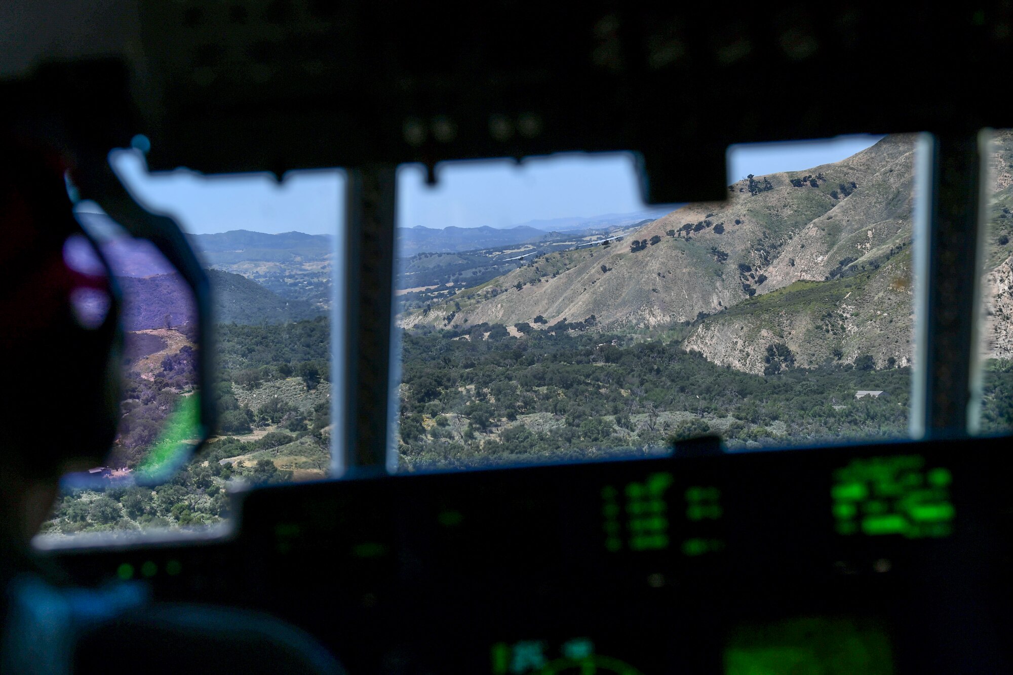 A view from inside the cockpit or flight-deck from the third chair of a military California Air National Guard C-130J Super Hercules aircraft. Inside the cockpit the back of the number one pilot can be seen as he stares into his heads-up-device as he closely follows two additional C-130J Super Hercules Aircraft in a green valley surrounded by a mountainous terrain.